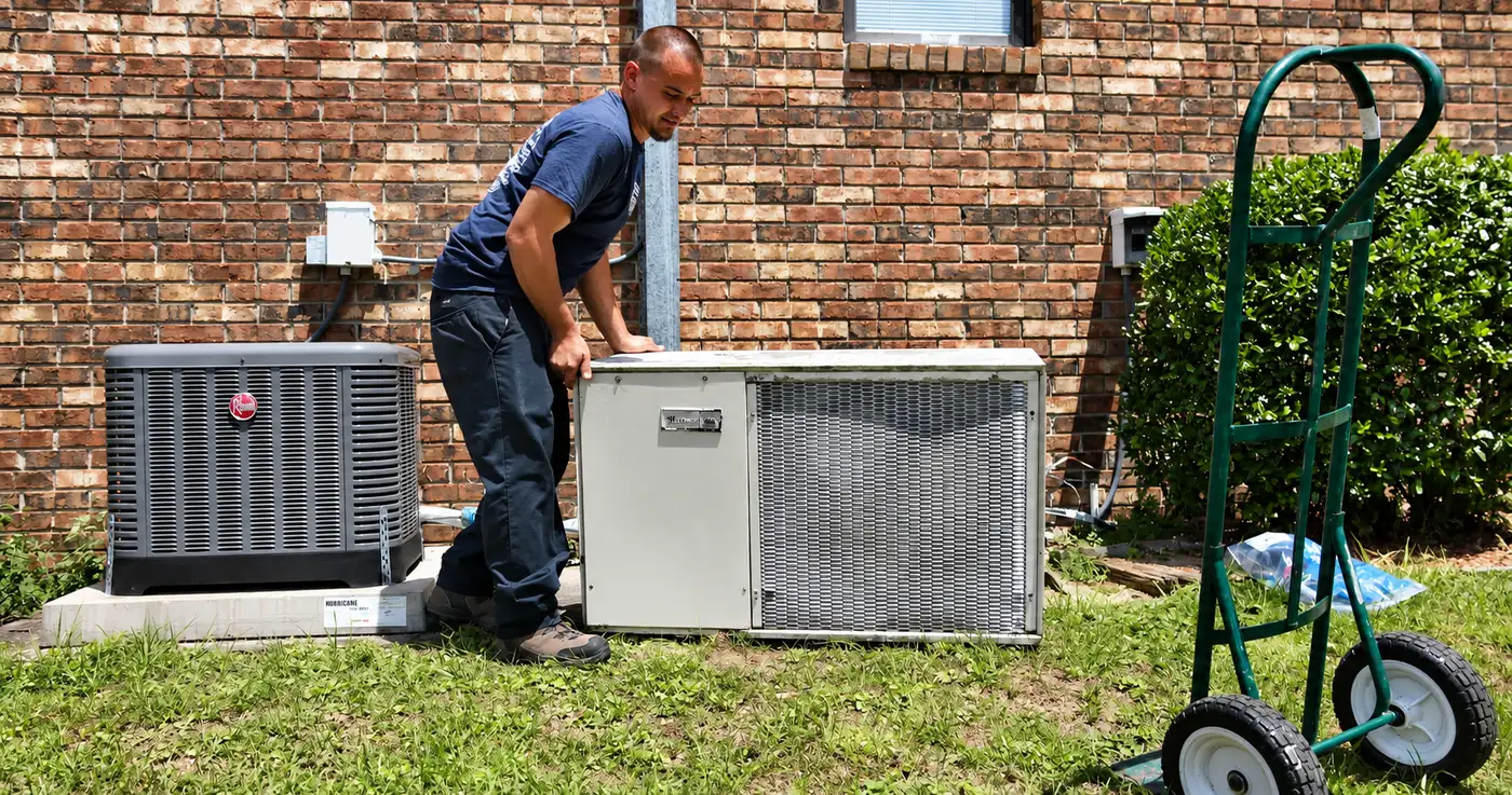 A technician removing an older outdoor air conditioning unit with its replacement condenser waiting alongside