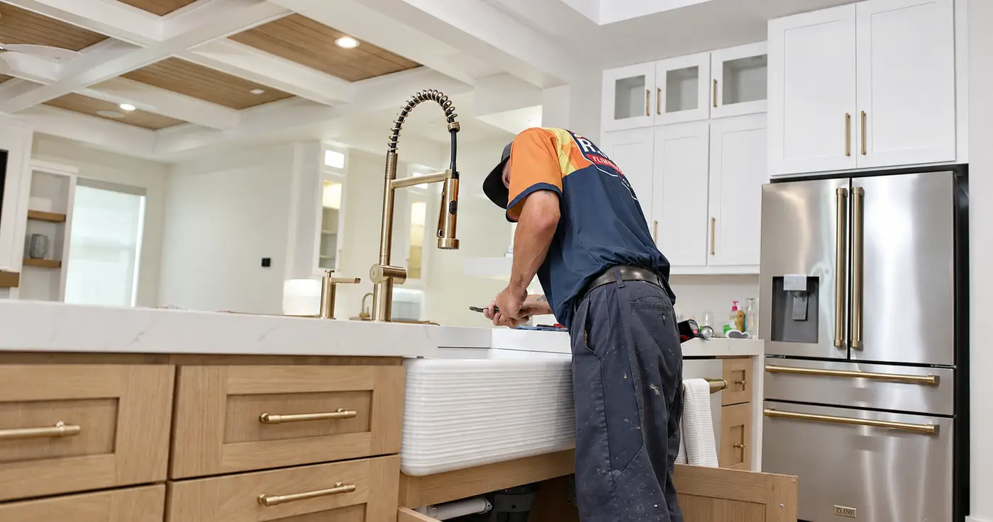 A technician working at a large kitchen sink and brass faucet with the cabinet below open