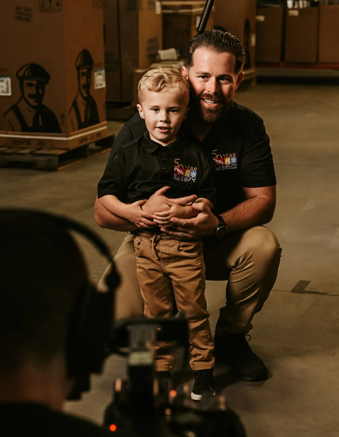 Tanner Kielty in an R.J. Kielty 50-year polo, kneeling beside a young boy in the company warehouse